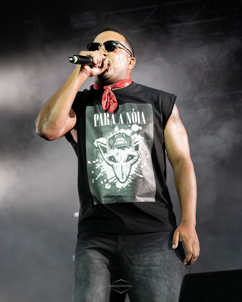 Male rapper performing on stage, wearing sunglasses, red bandana, and black sleeveless graphic tee, singing into microphone.