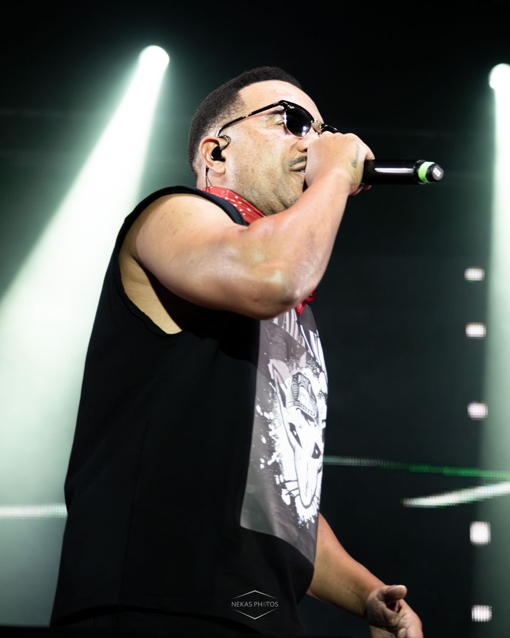 Male rapper performing on stage with microphone, wearing black sleeveless shirt and sunglasses under dramatic green stage lighting.
