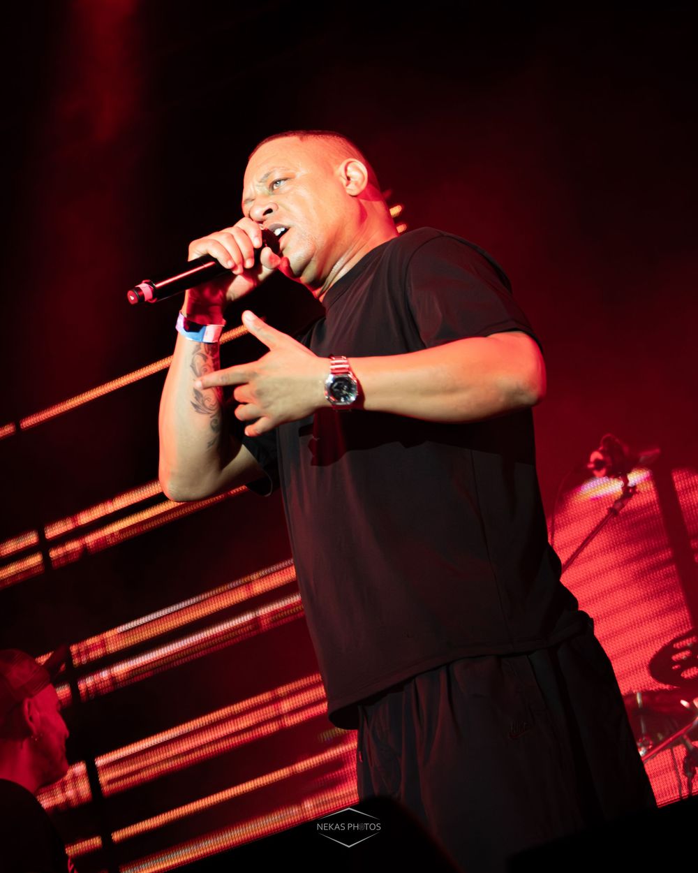 Bald male rapper performing on stage holding microphone under red stage lighting wearing black t-shirt and red watch.