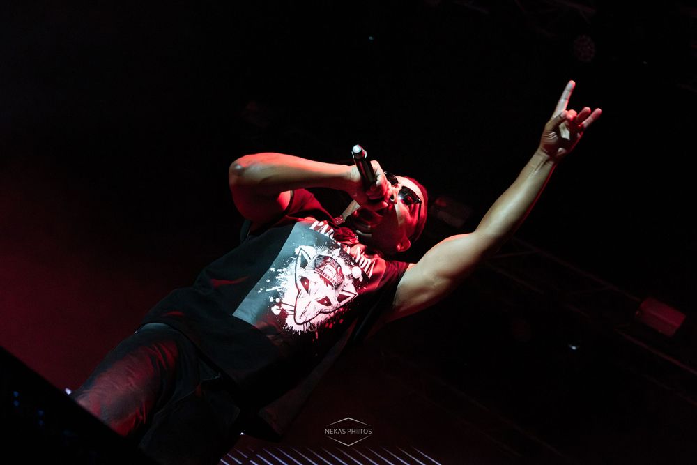 Male rock singer performing on stage, making devil horns gesture while holding microphone under dramatic red lighting.