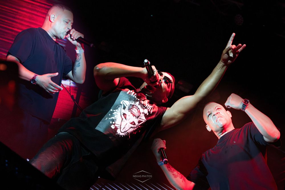 Three performers energetically rapping on a dark red-lit stage during a live hip-hop concert performance.