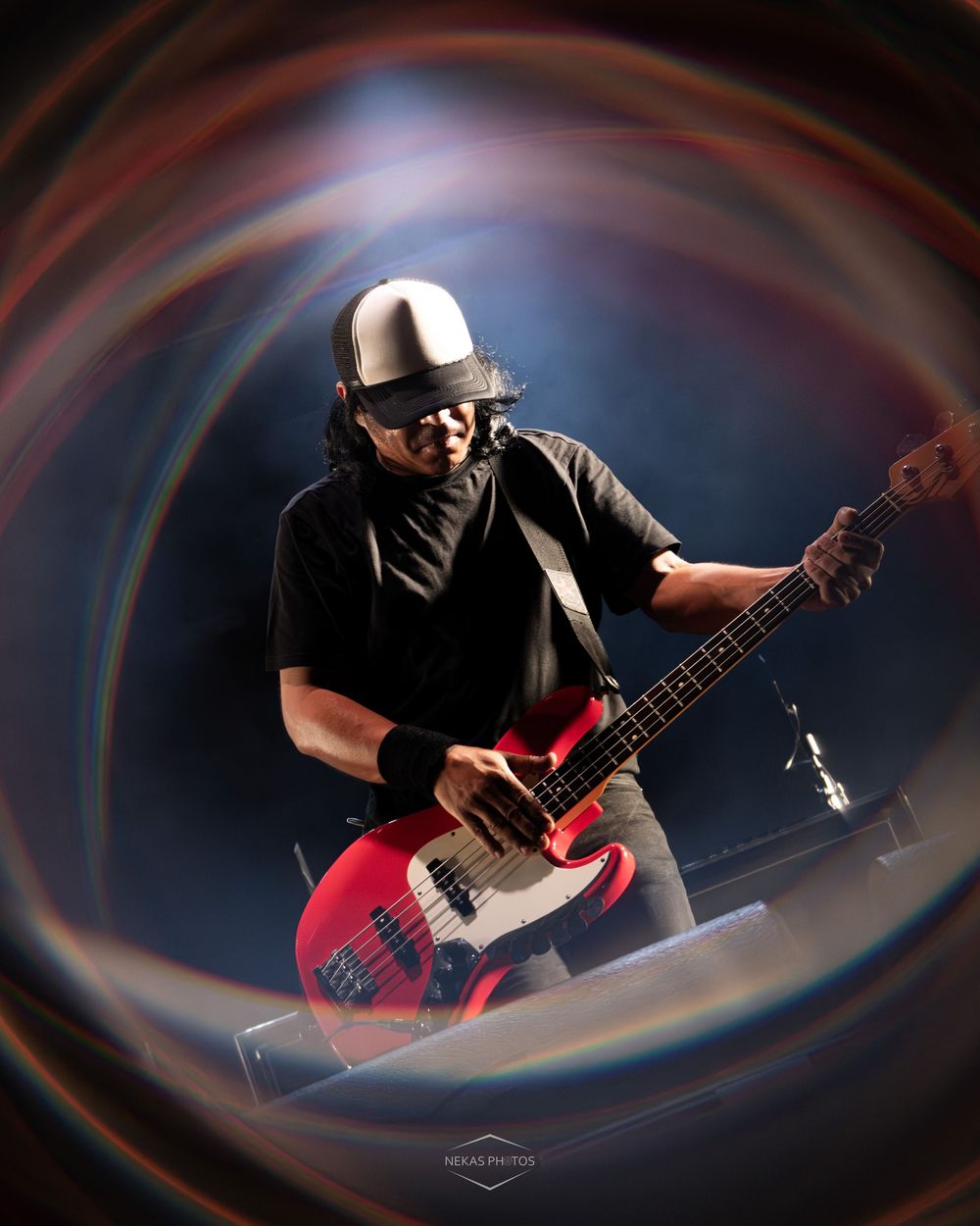 Guitarist playing red bass guitar on stage with swirling colorful light effect and silver bucket hat.