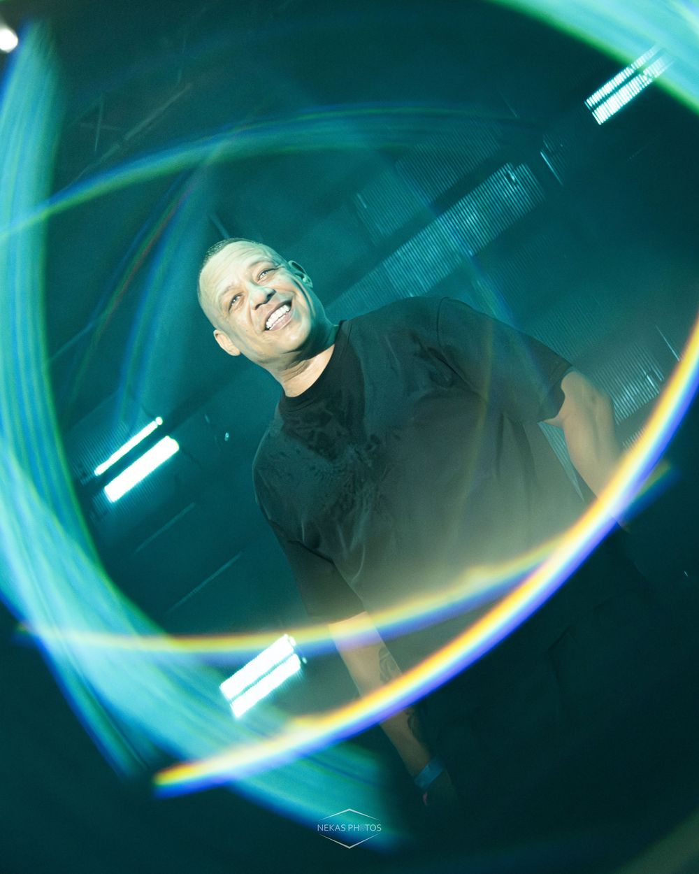 Smiling bald man photographed through a circular lens flare with teal neon lights creating a vibrant, artistic effect.