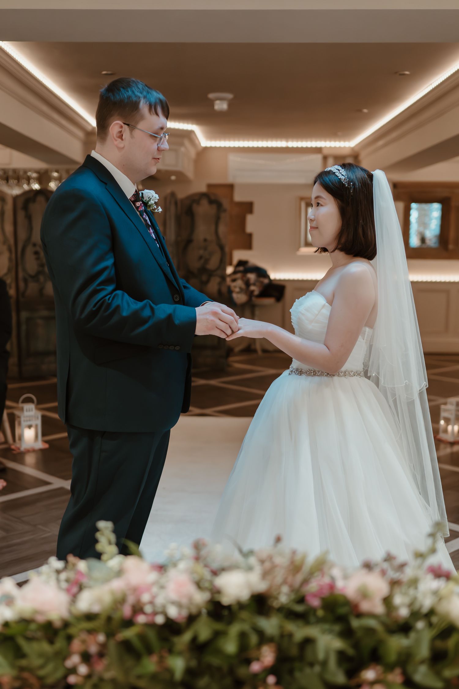 Groom placing wedding ring on bride's finger during elegant indoor ceremony, bride in white gown with veil.