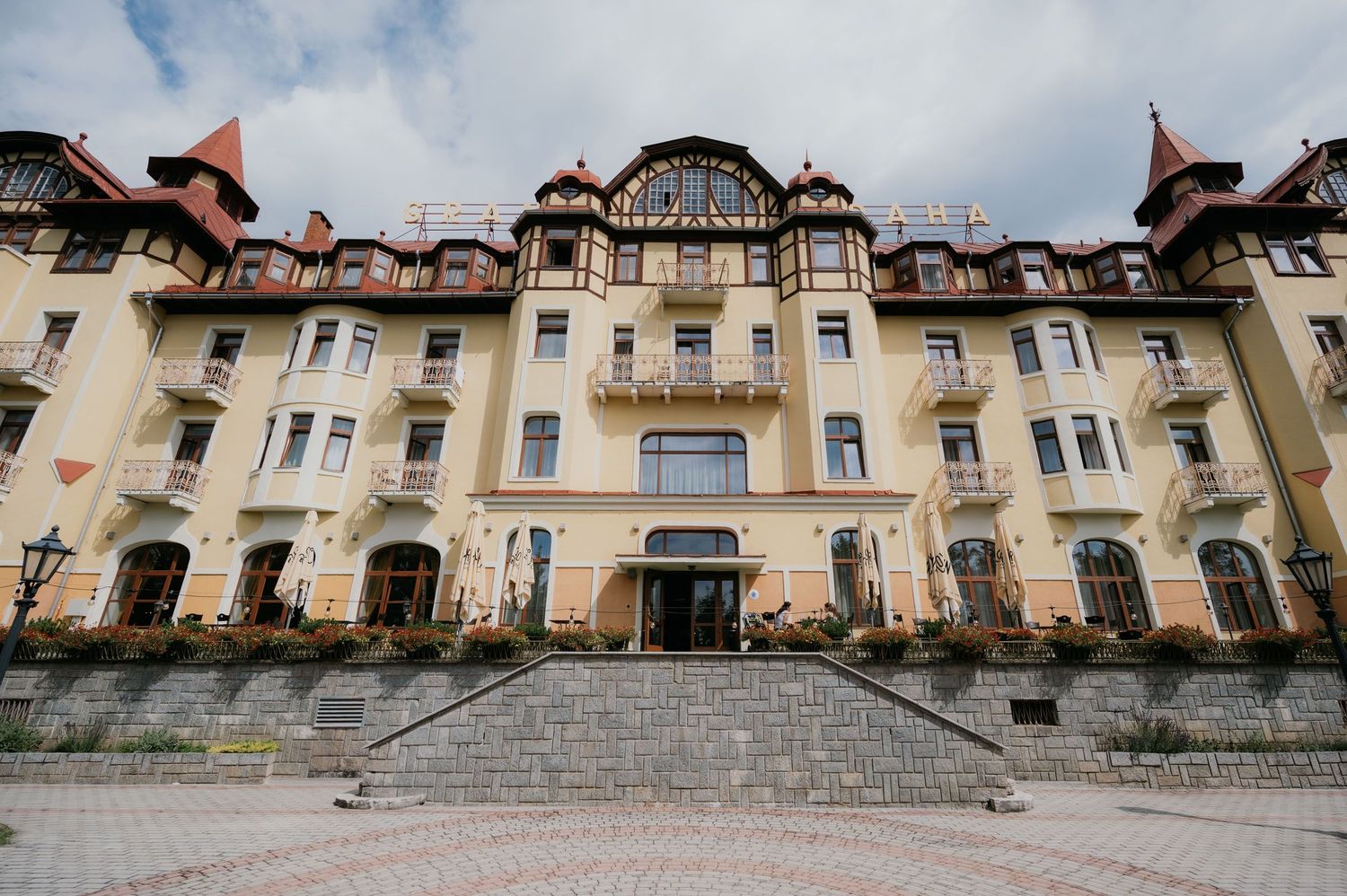 Grand yellow historic hotel with ornate Art Nouveau facade, arched entrance, balconies, and cobblestone plaza under cloudy sky.