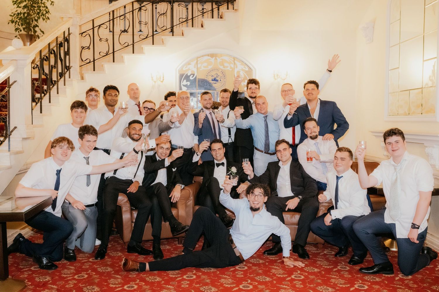 Large group of men in formal attire posing together on a red carpet staircase at a wedding celebration.