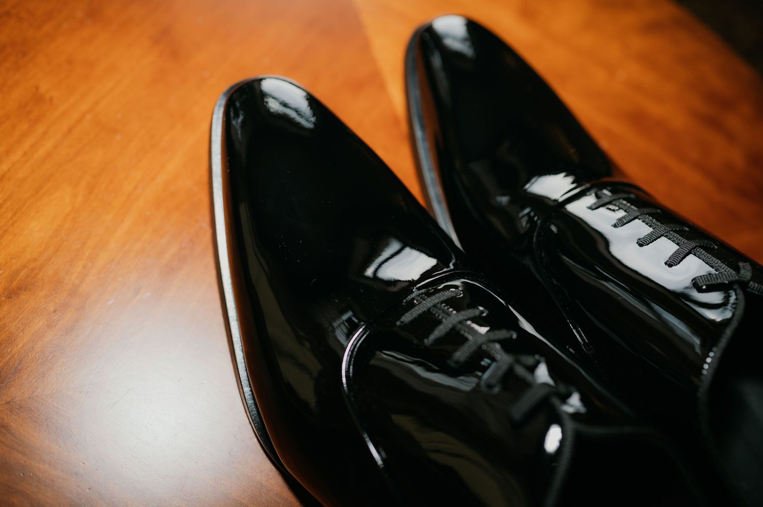 Shiny black patent leather oxford dress shoes with laces on a wooden surface.
