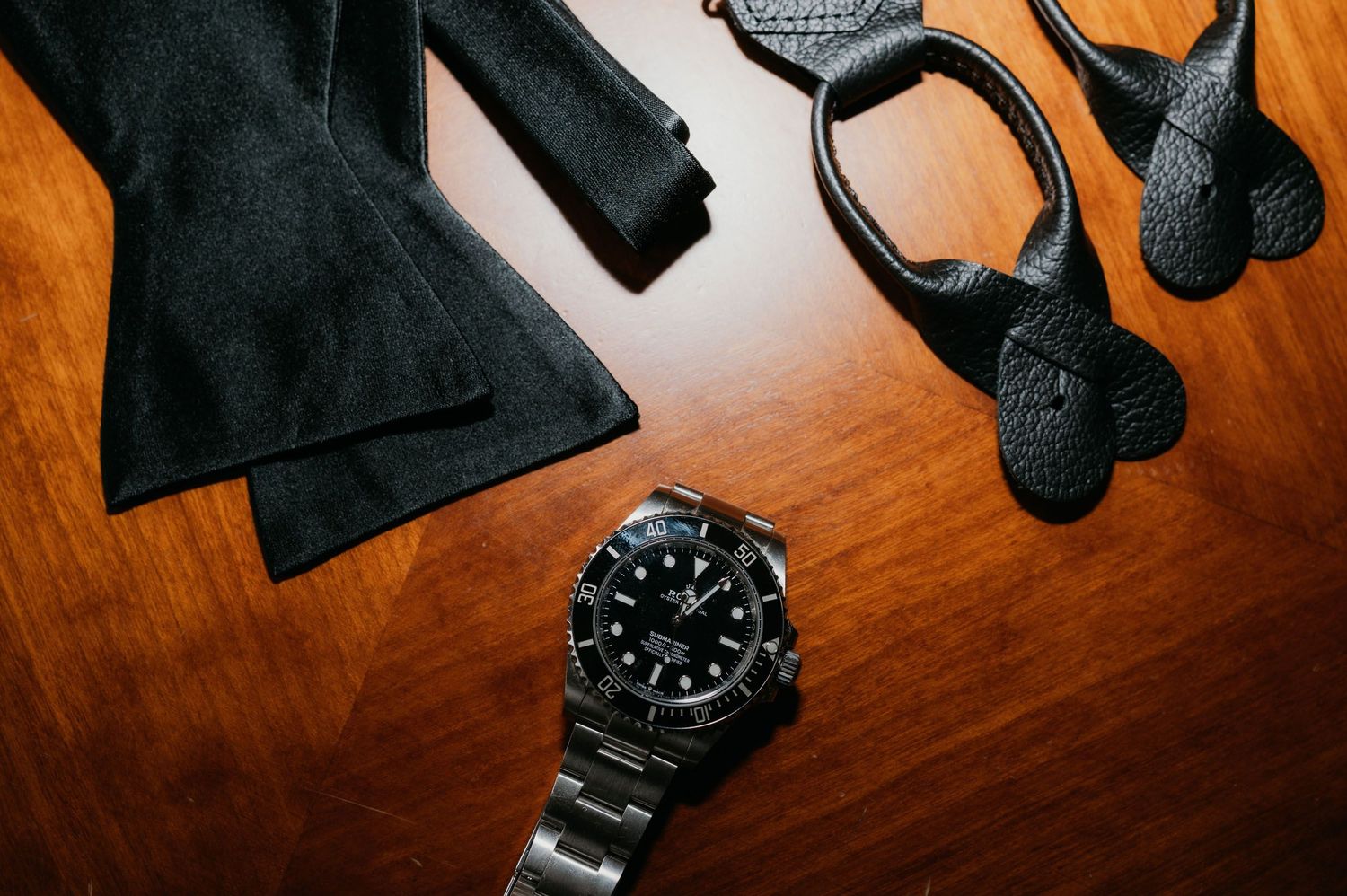 Rolex Submariner watch with black dial beside a bow tie and leather accessories on a wooden surface.