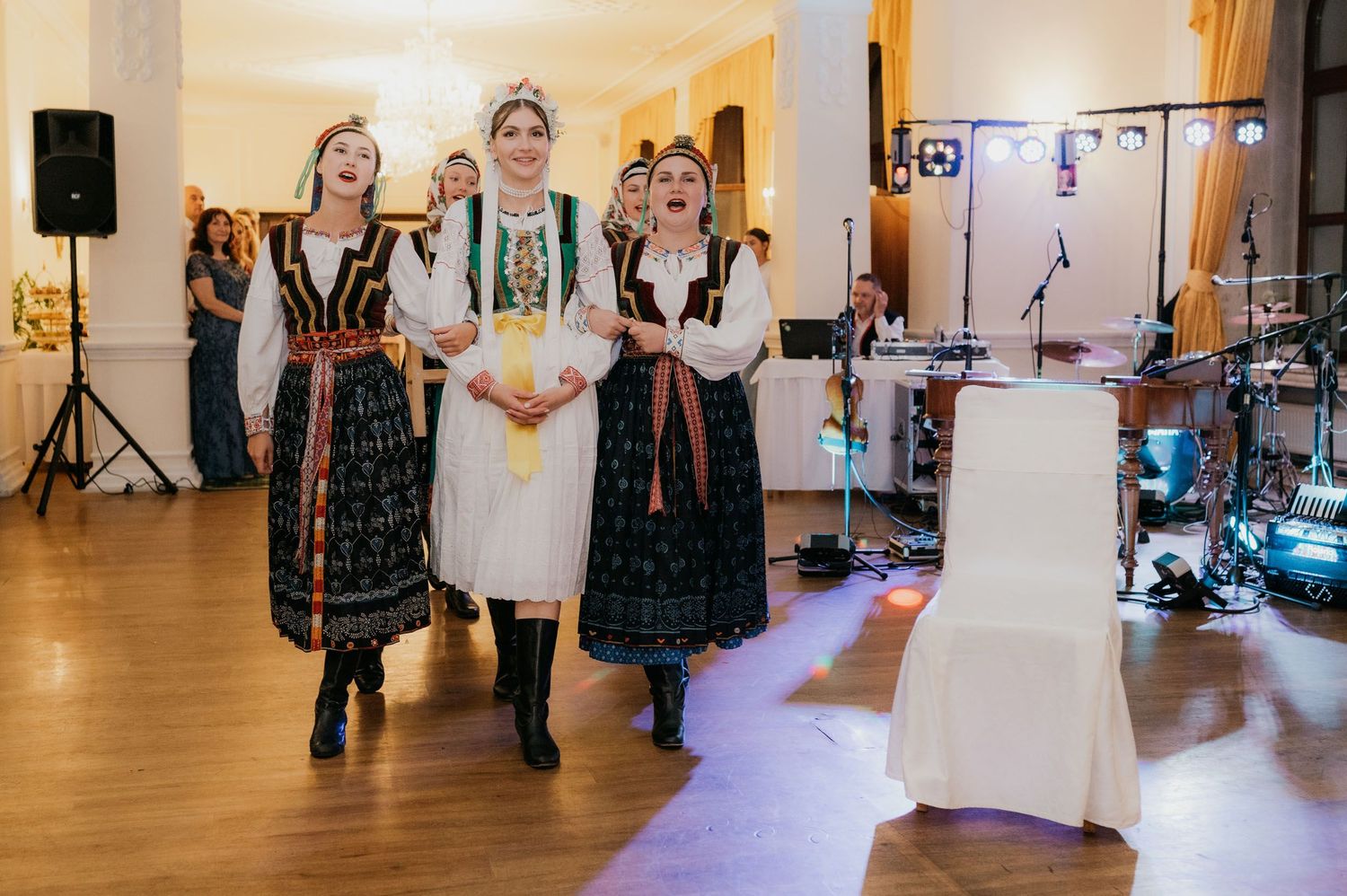 Three women in traditional Eastern European folk costumes singing at a wedding reception.