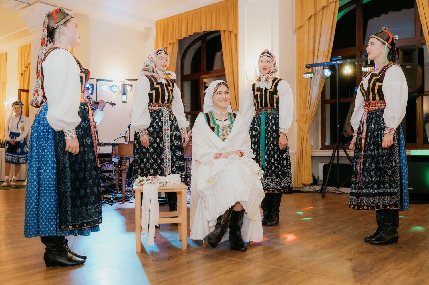 Women in traditional folk costumes perform a cultural dance ceremony in an ornate hall.