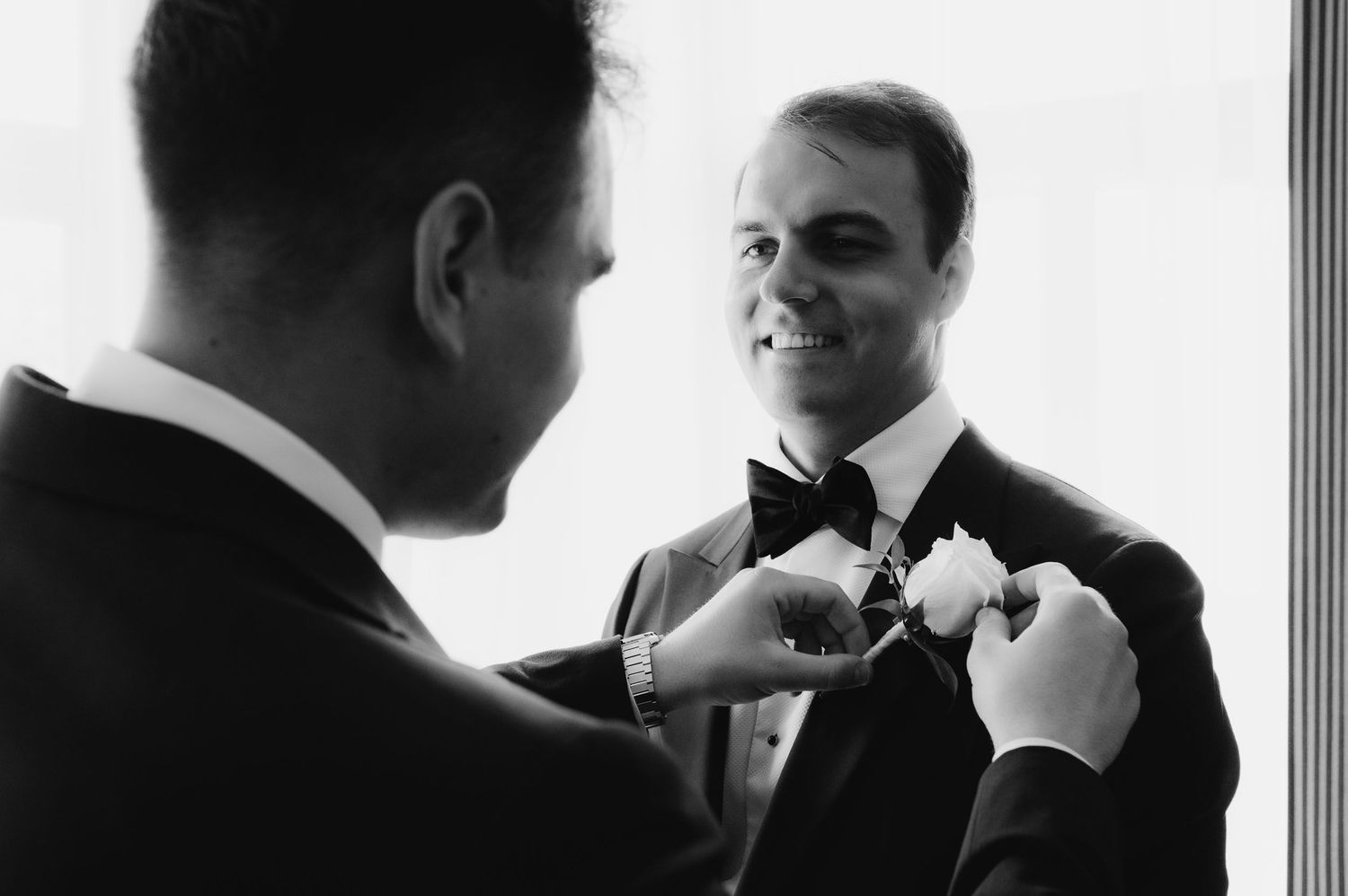 Black and white photo of groom smiling as best man pins boutonniere on tuxedo lapel before wedding ceremony.