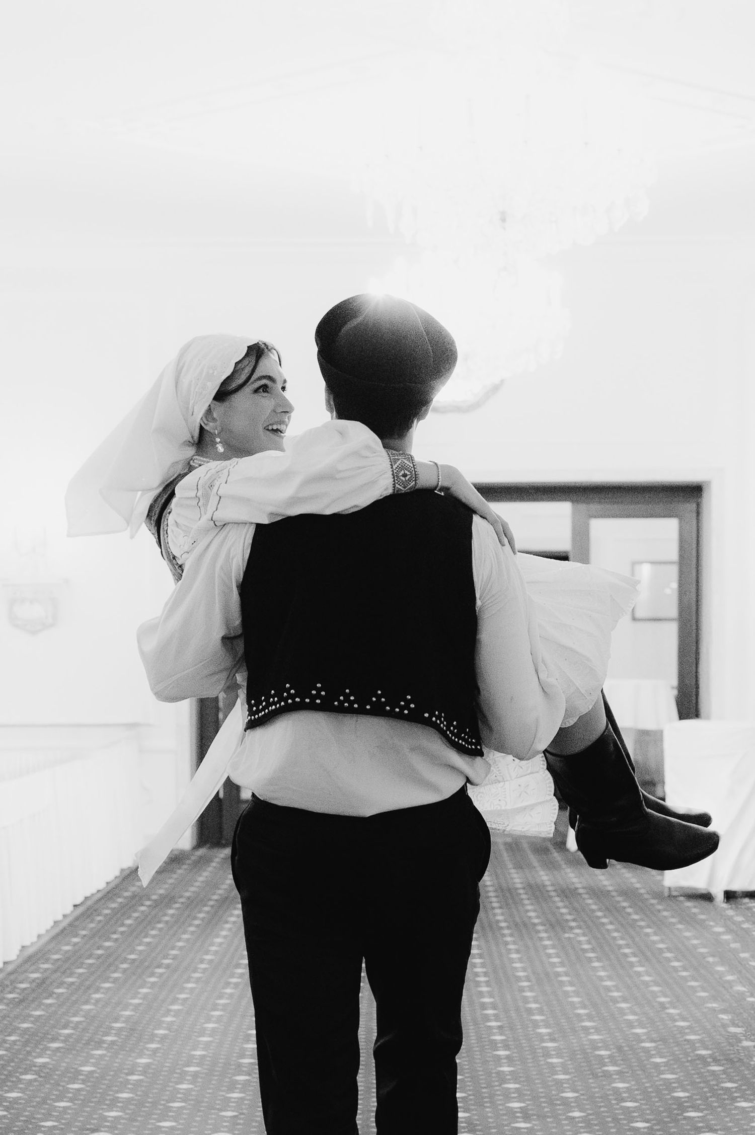 Black and white photo of a groom carrying his smiling bride in a wedding venue.