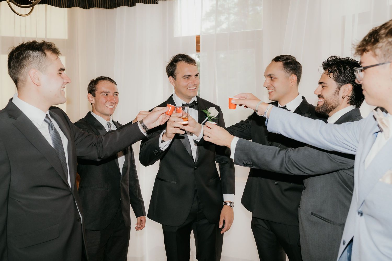Groom and groomsmen in suits toasting with mini bottles at a wedding celebration indoors.