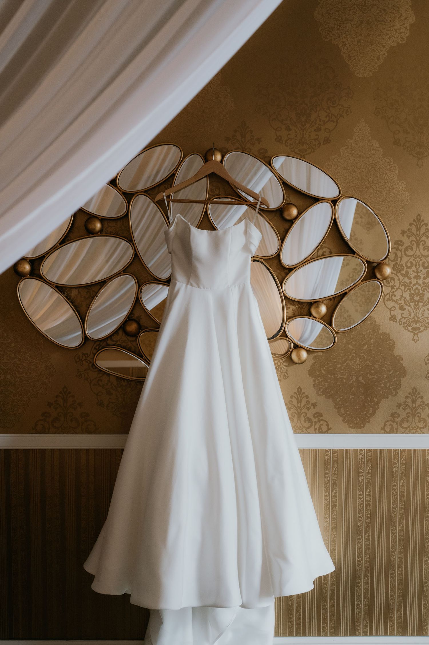 Elegant white A-line wedding dress hanging on a decorative gold mirror against a gold damask wall.