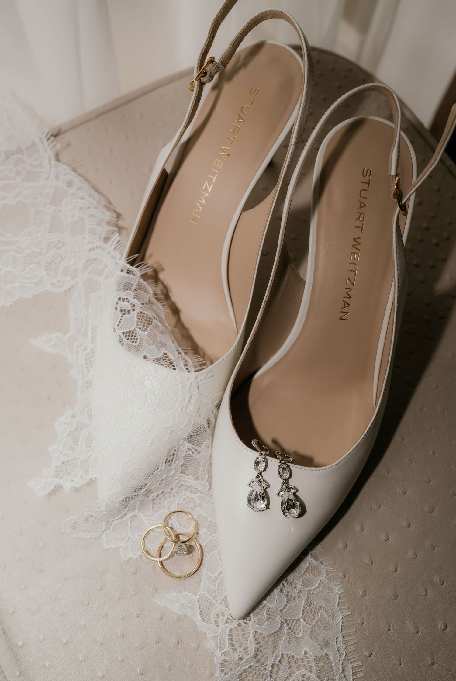 Stuart Weitzman white slingback heels with crystal earrings and gold wedding bands on lace fabric.