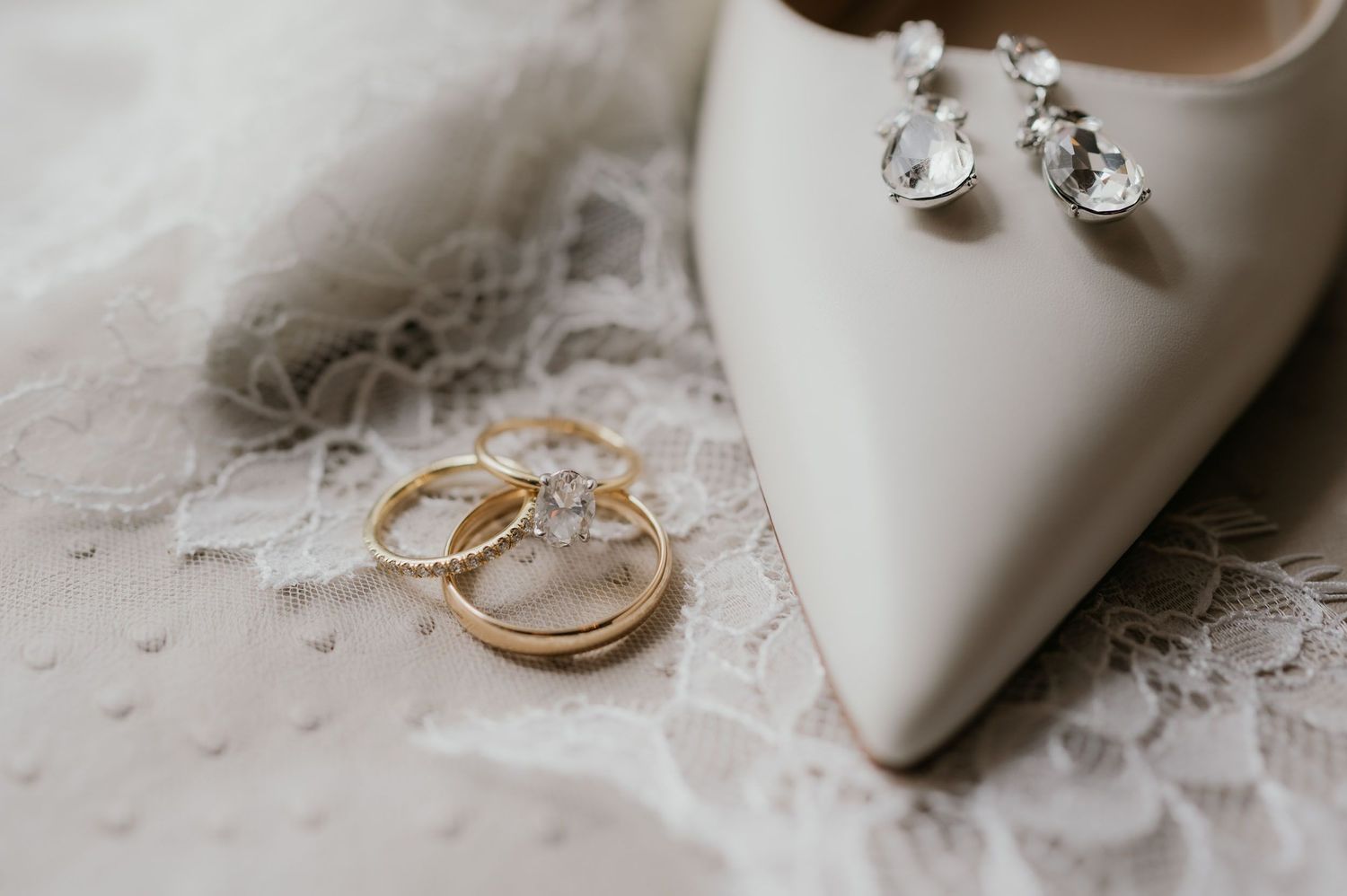 Gold wedding bands rest beside a white bridal heel and crystal drop earrings on delicate lace fabric.
