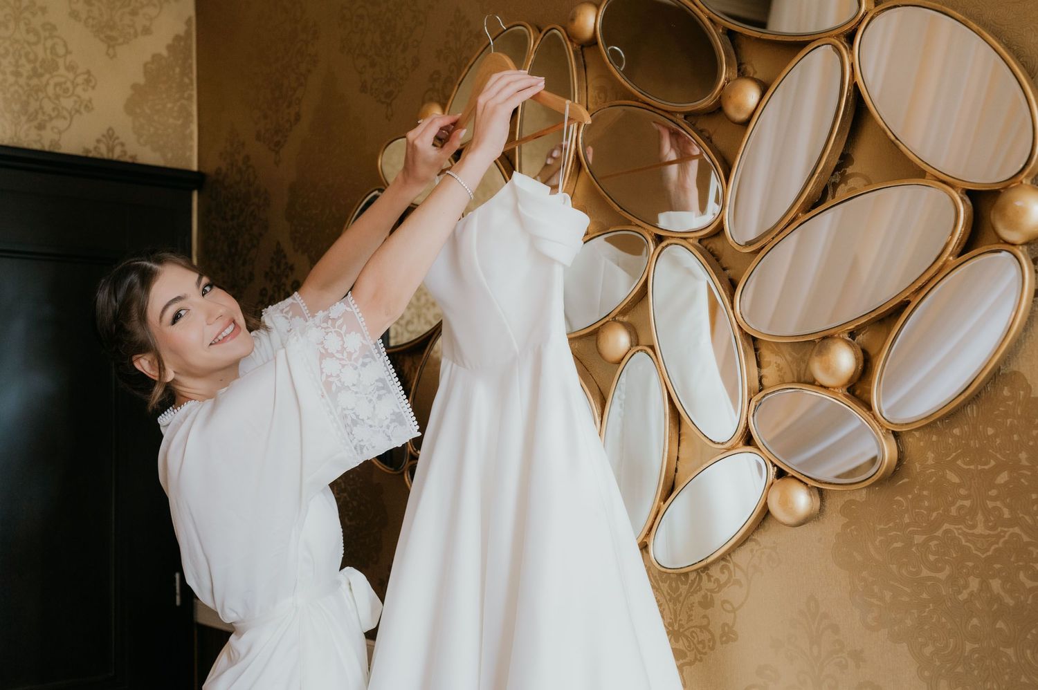 Bride in white robe holding wedding dress on hanger in front of decorative gold mirror wall.