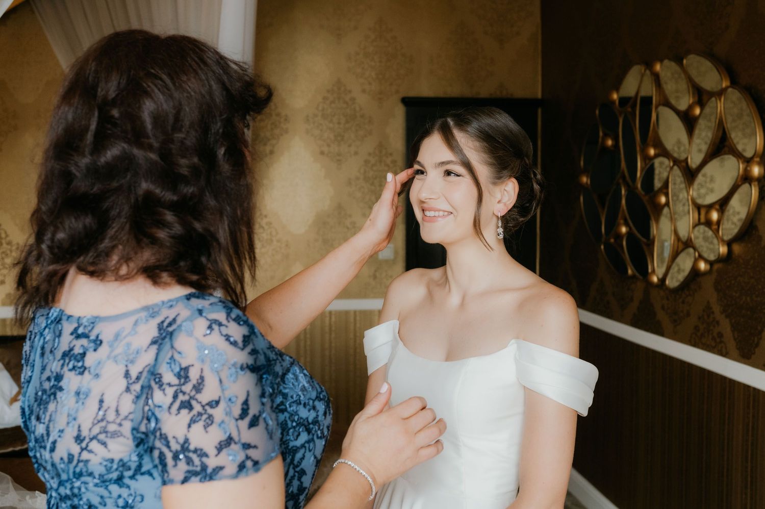 A woman in a blue dress adjusts a smiling bride's hair as she prepares for her wedding day.