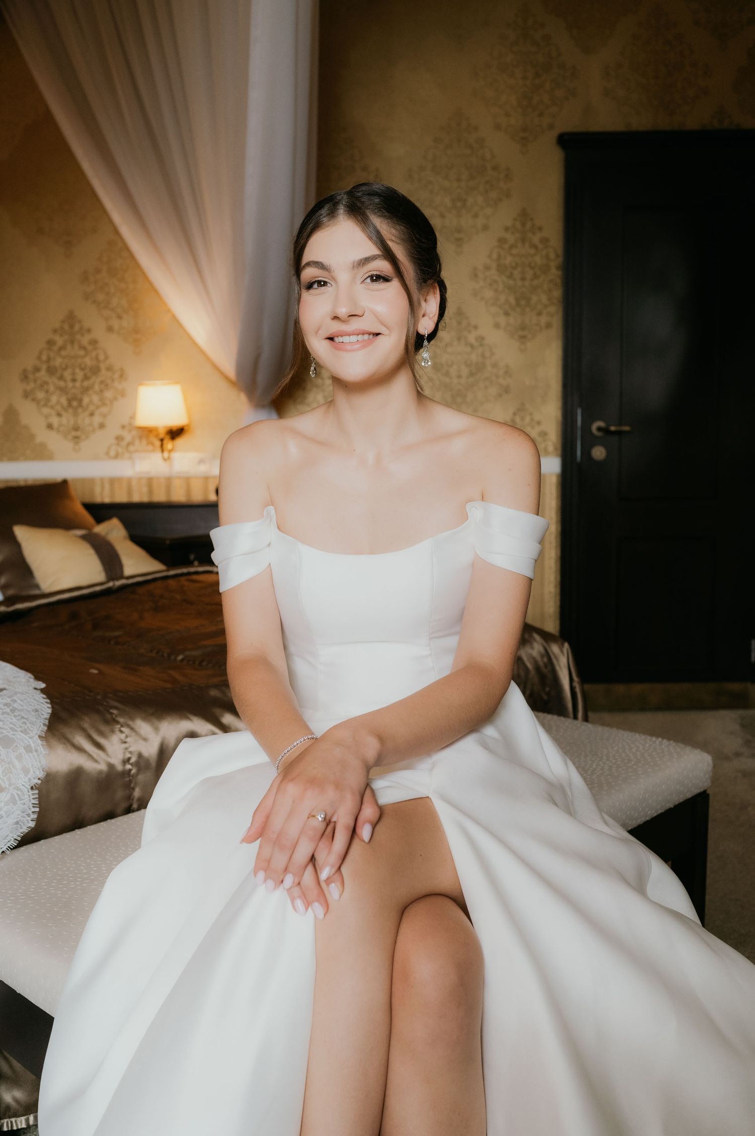 Smiling bride in white off-shoulder wedding dress sitting elegantly in a decorated hotel room.