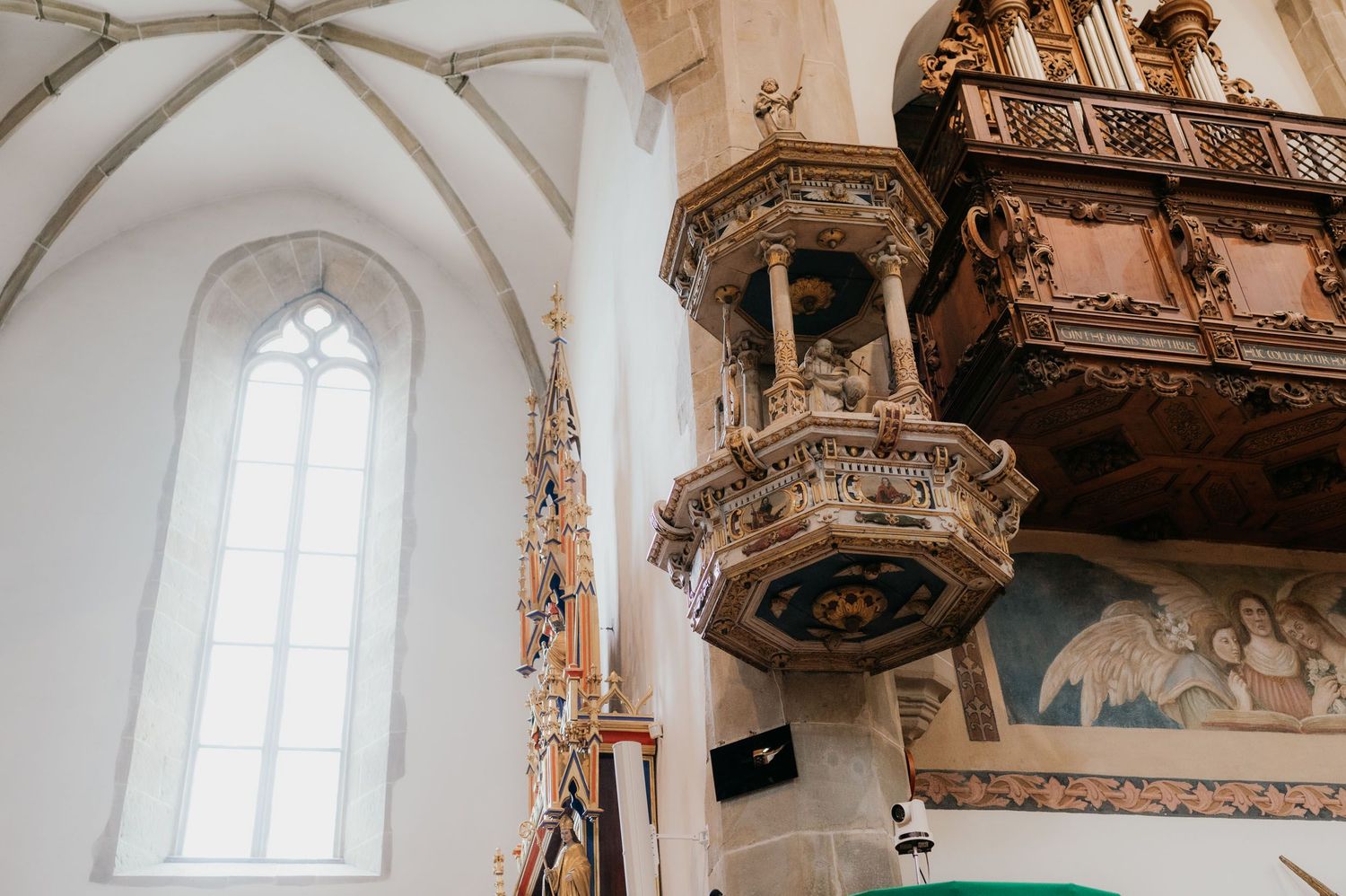 Ornate carved wooden pulpit inside a Gothic church with white vaulted ceiling and stained glass.