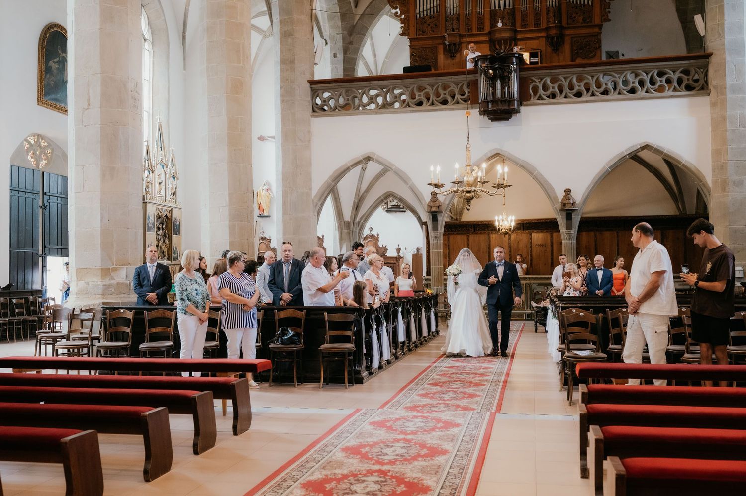 Wedding ceremony inside a Gothic church with guests standing along red carpet aisle as couple exchanges vows.