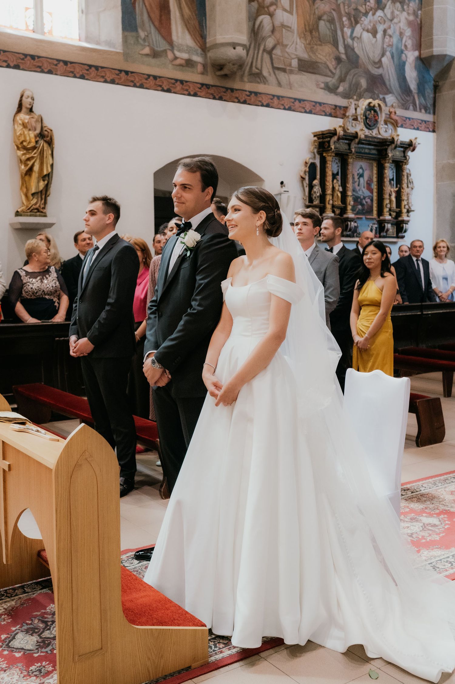 Bride and groom stand at the altar during a church wedding ceremony, surrounded by guests in an ornate chapel.