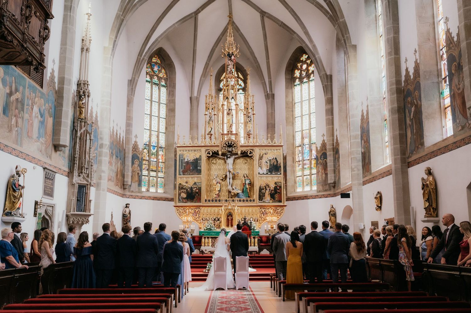 Wedding ceremony in a stunning Gothic church with ornate altar, frescoes, and guests gathered in pews.