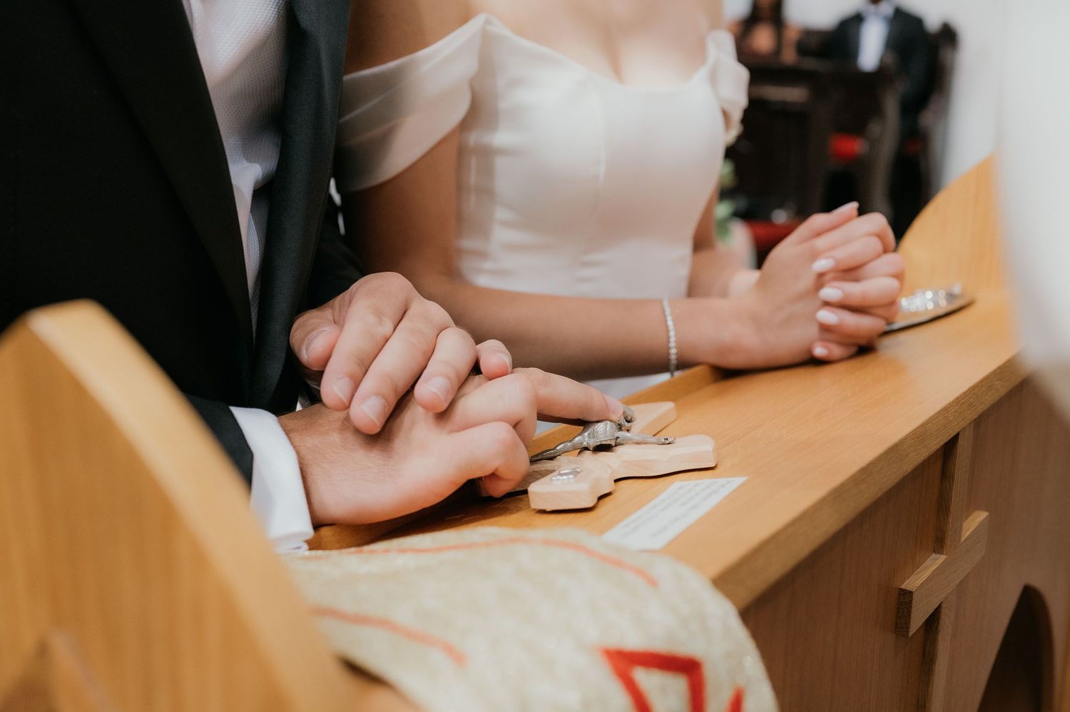 Bride and groom holding hands at wedding ceremony altar, exchanging rings on wooden church pew.