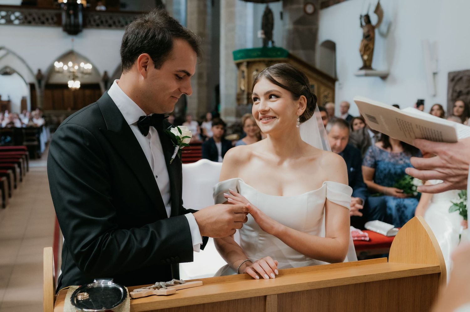 Bride and groom exchanging rings at church altar during wedding ceremony, bride smiling in off-shoulder white gown.