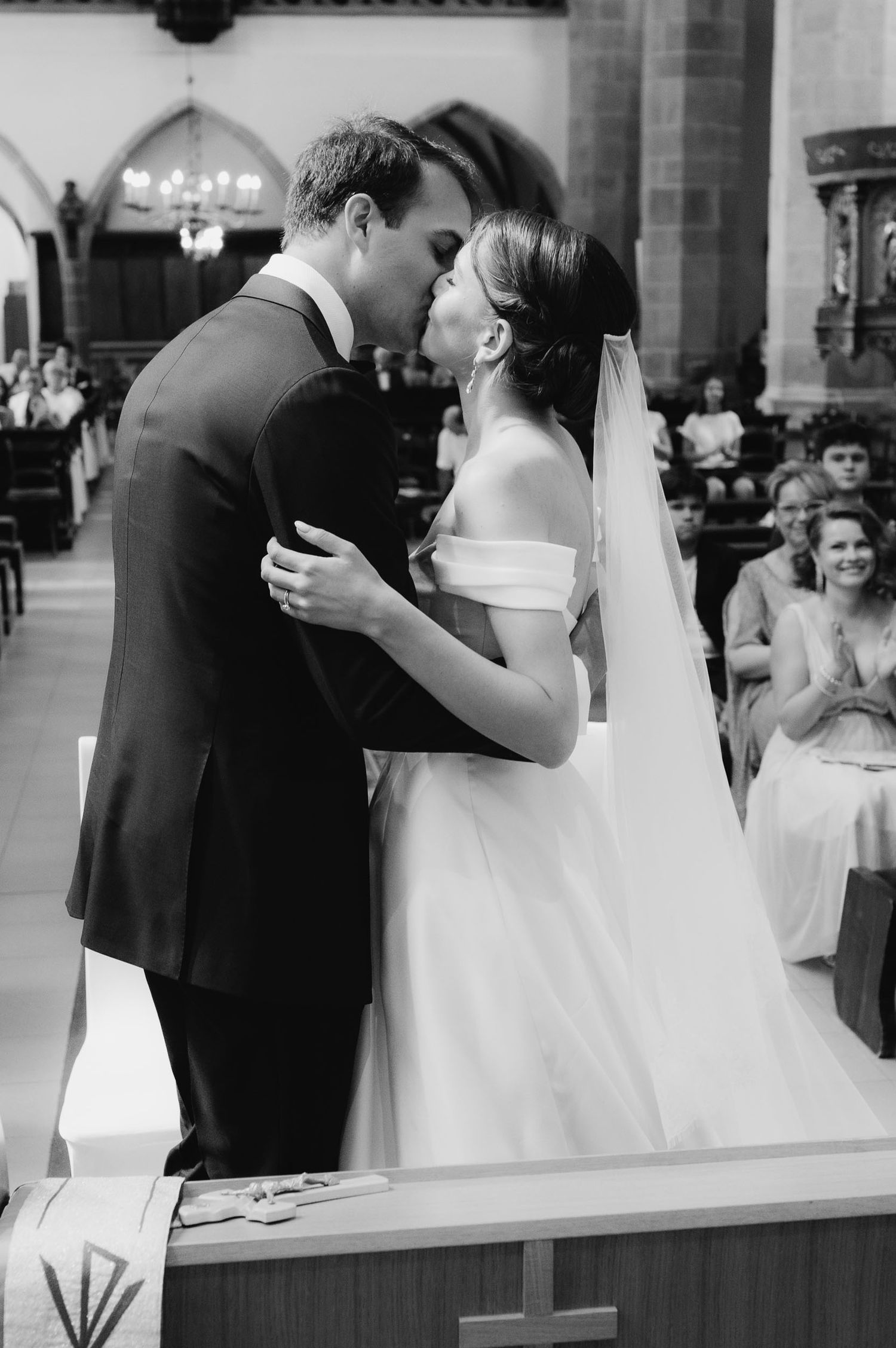 Black and white photo of a bride and groom sharing their first kiss at the altar during a church wedding ceremony.