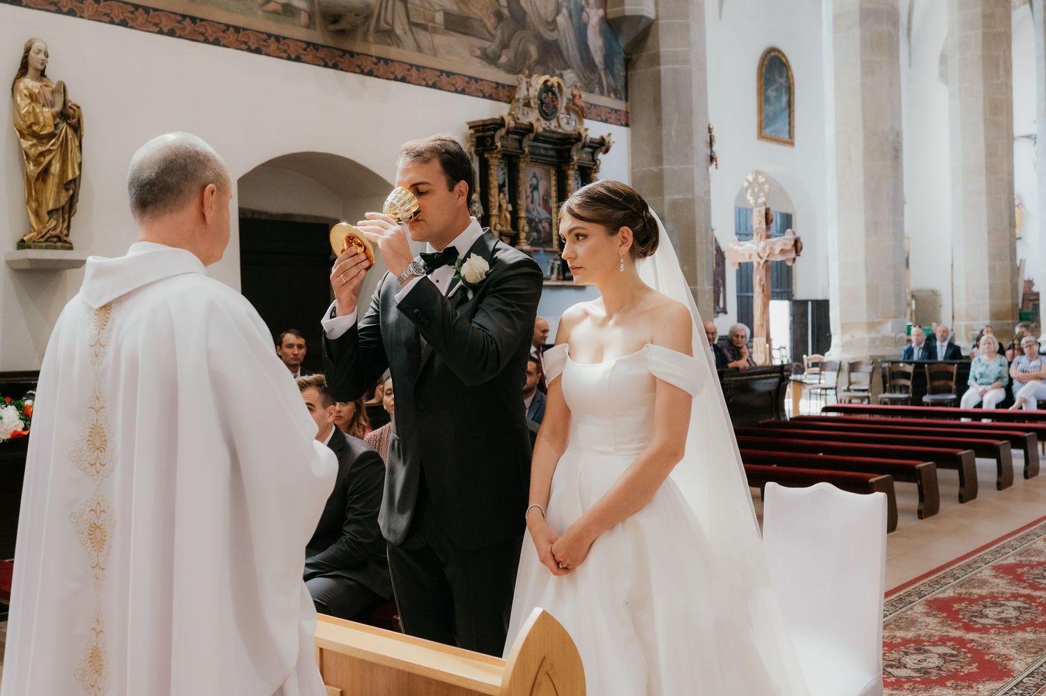 Groom drinking from chalice during Catholic wedding ceremony at church altar with bride in white gown.