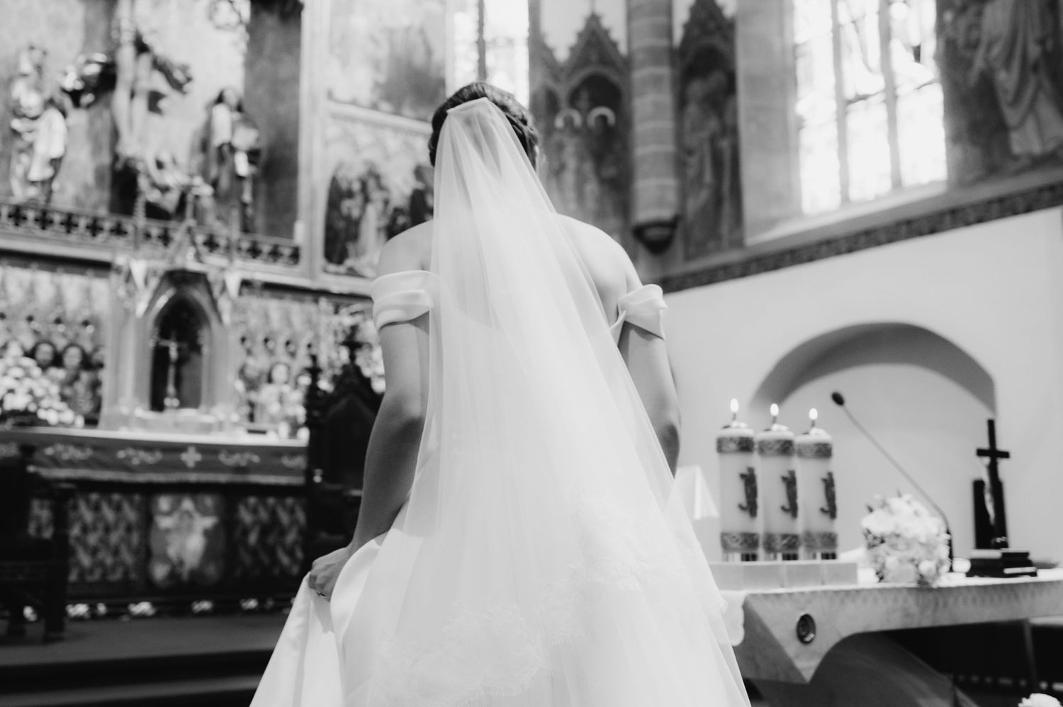 Bride in white wedding gown and veil stands at church altar during ceremony, black and white photo.