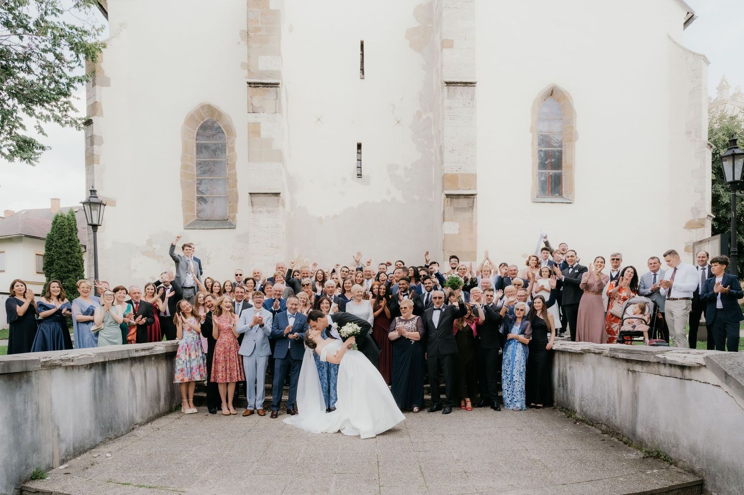 Wedding couple kissing in front of a church surrounded by cheering guests at an outdoor group photo.