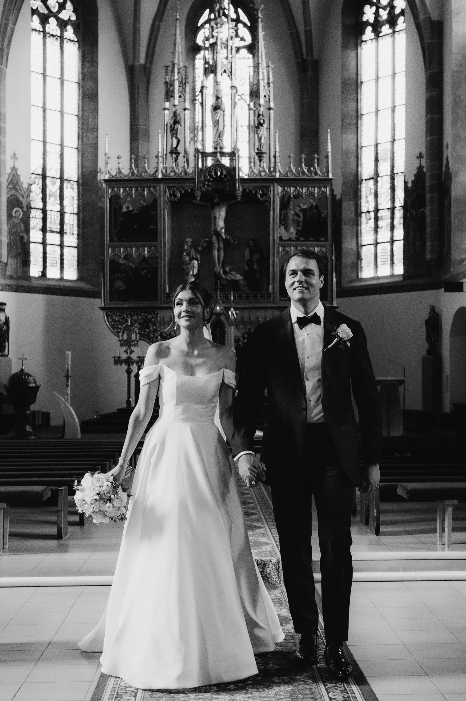 Black and white photo of a bride and groom holding hands at the altar of an ornate church after their wedding ceremony.