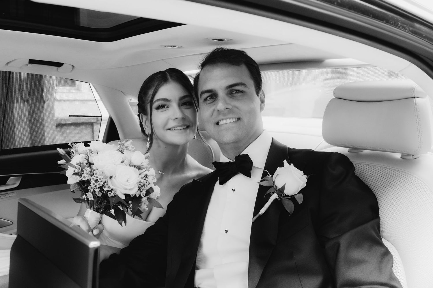 Black and white photo of a bride and groom smiling in a car, bride holding a bouquet, groom in a tuxedo with boutonniere.