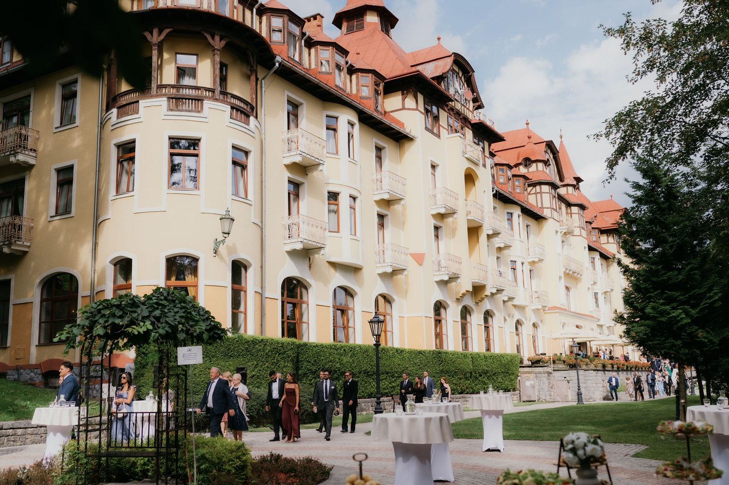 Elegant yellow historic hotel building with ornate architecture, surrounded by lush gardens and guests at an outdoor event.