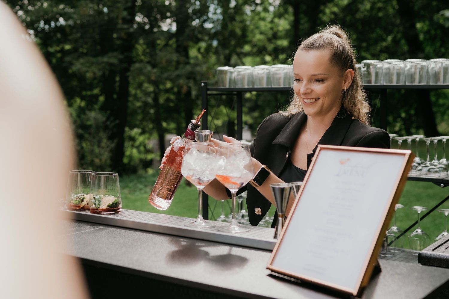 Smiling female bartender pouring pink cocktails at an outdoor bar during an elegant event.