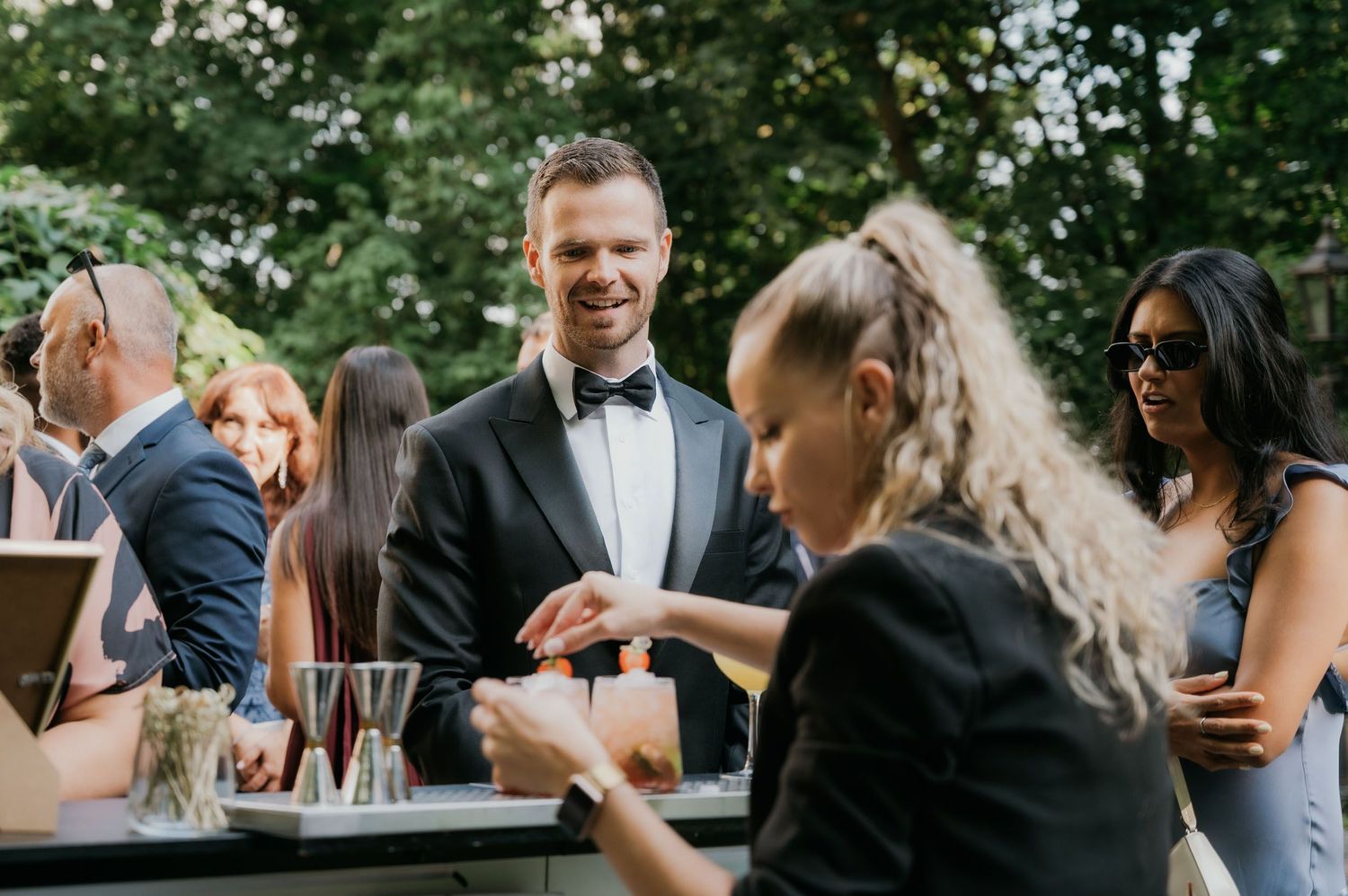 Man in tuxedo smiling at outdoor wedding reception bar, guests socializing in background.