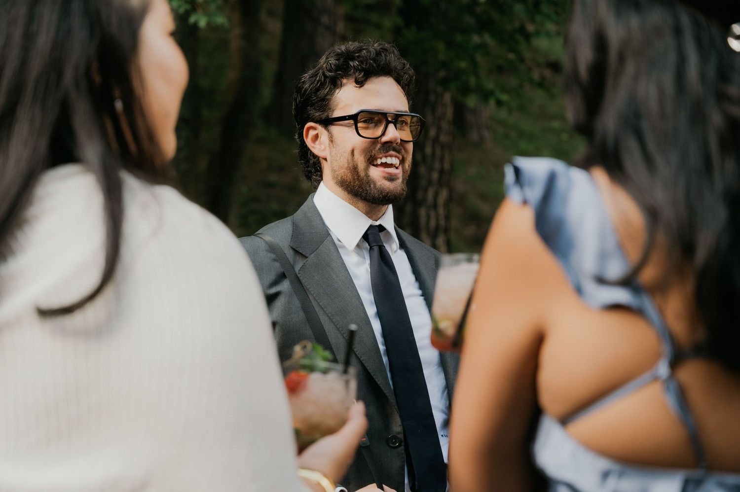 Smiling man in gray suit and black tie laughing with guests at an outdoor wedding reception.