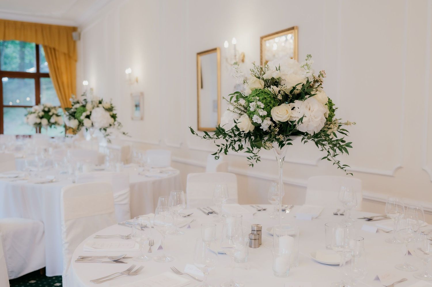 Elegant wedding reception hall with white floral centerpieces, white tablecloths, and gold curtains in a bright, airy venue.