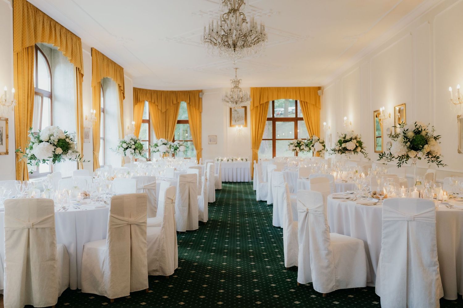 Elegant wedding reception hall with white chair covers, round tables, green carpet, gold curtains, and floral centerpieces.