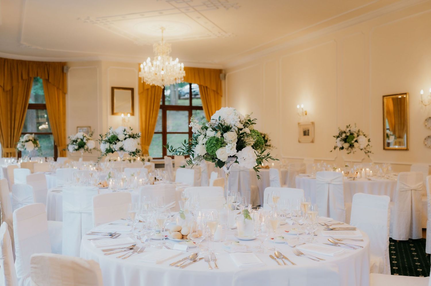 Elegant wedding reception hall with white round tables, floral centerpieces, chandelier, and gold curtains.