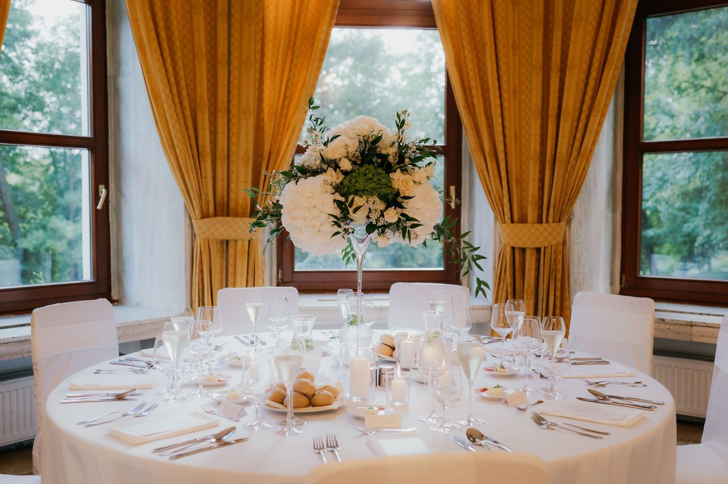 Intimate wedding table setting with white linens, candles, floral centerpiece, and golden drapes by window.