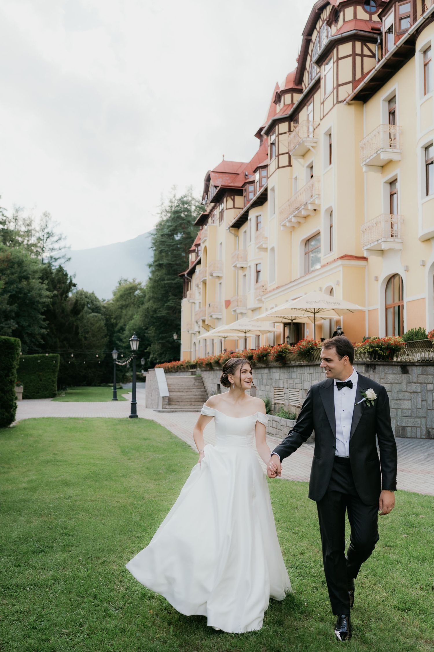 Bride and groom walk hand in hand on green lawn outside elegant European hotel on their wedding day.