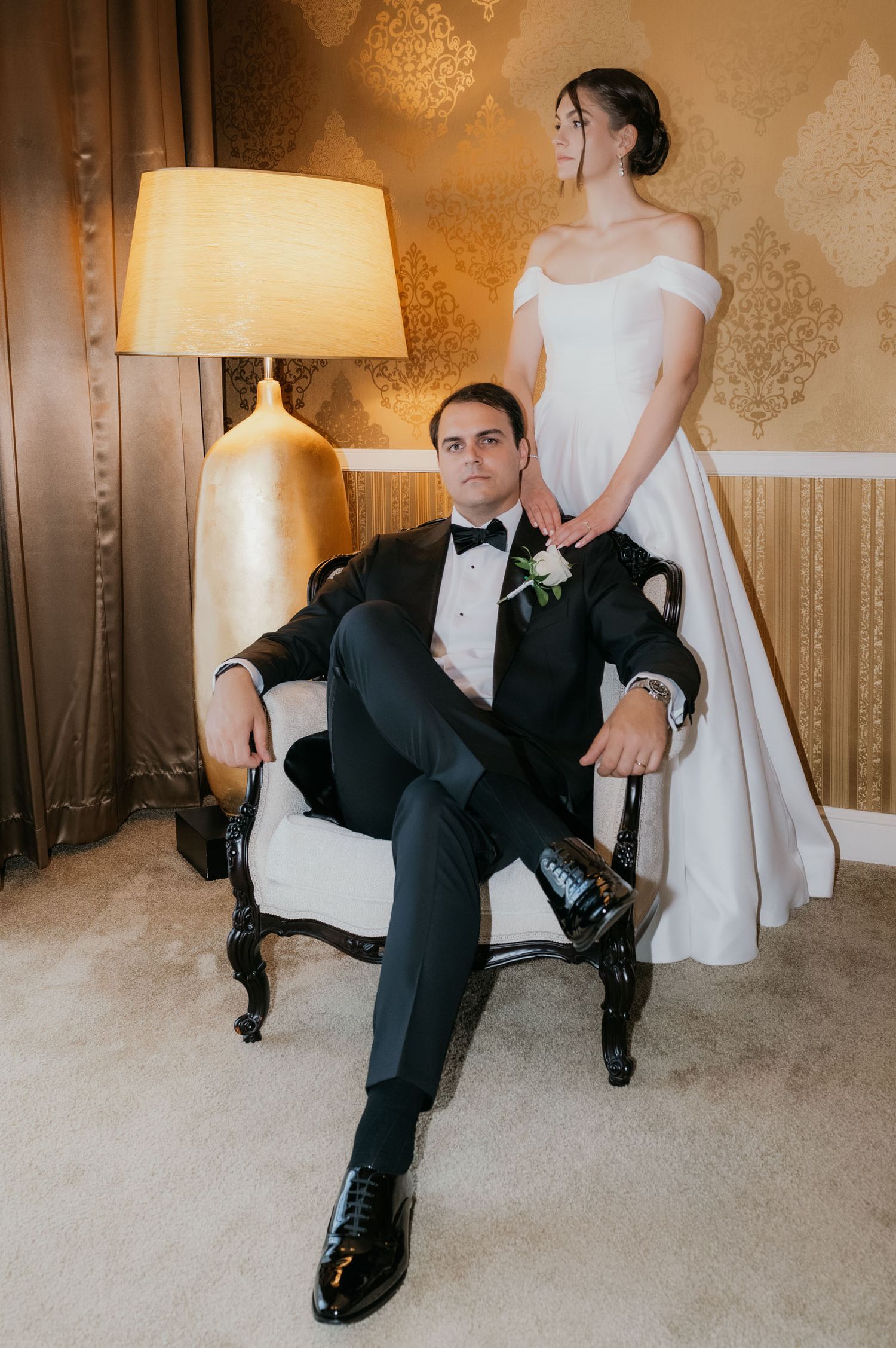Groom in black tuxedo sits in ornate chair while bride in off-shoulder white gown stands behind him in elegant room.