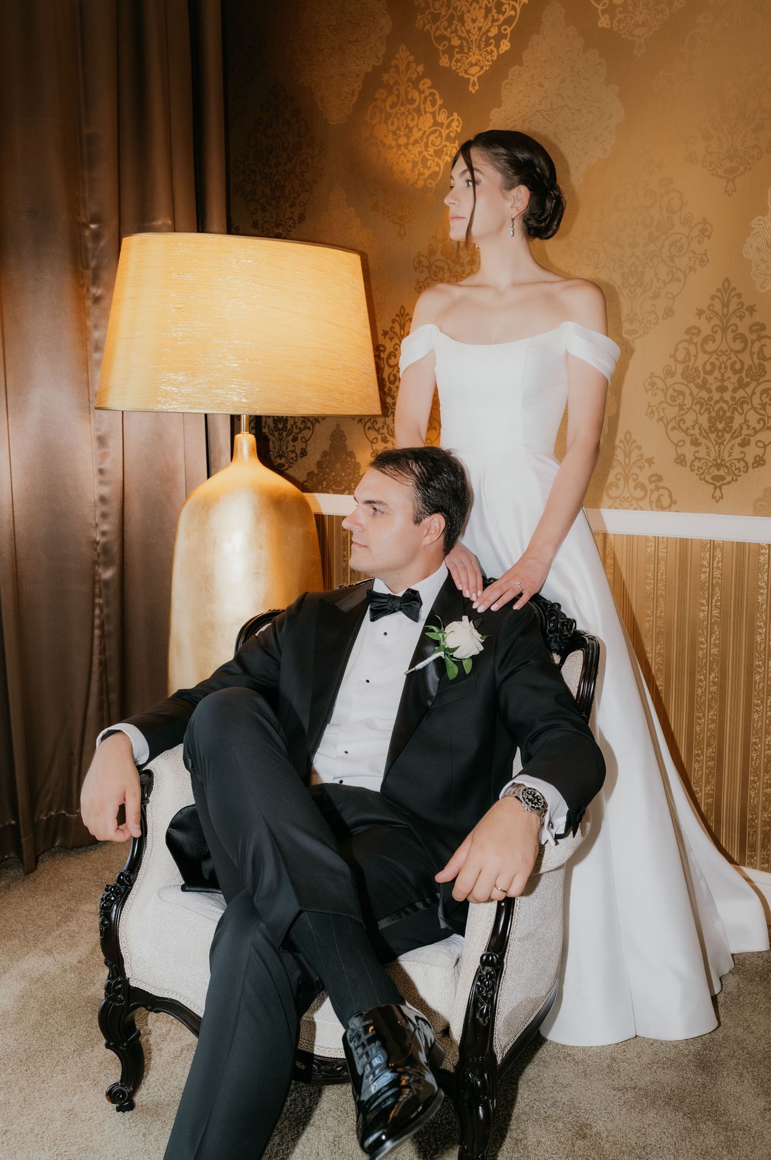 Elegant bride in off-shoulder gown stands behind groom in black tuxedo seated in ornate chair beside a gold lamp.