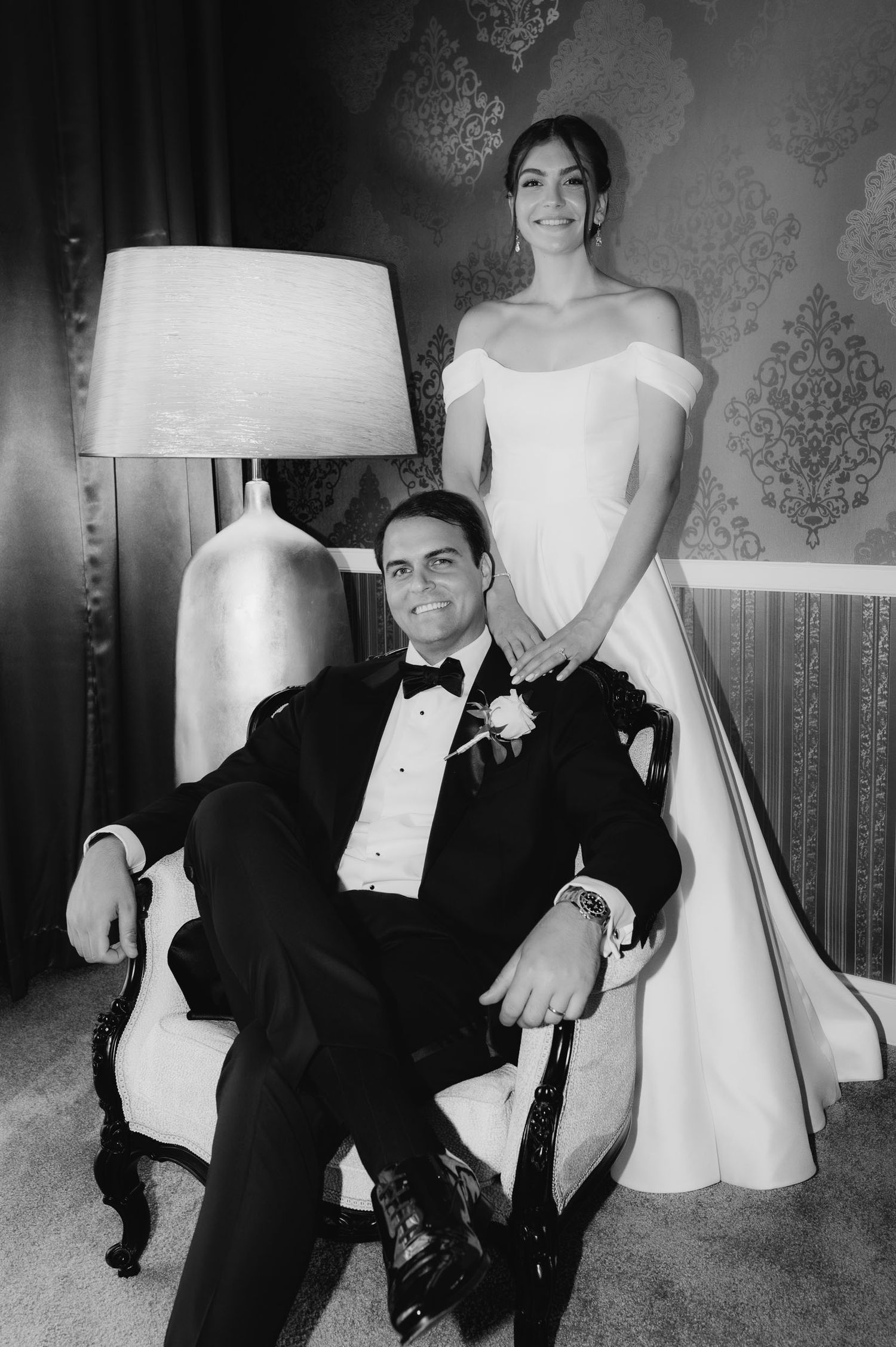 Black and white wedding portrait of bride in off-shoulder gown standing beside groom in tuxedo seated in ornate chair.