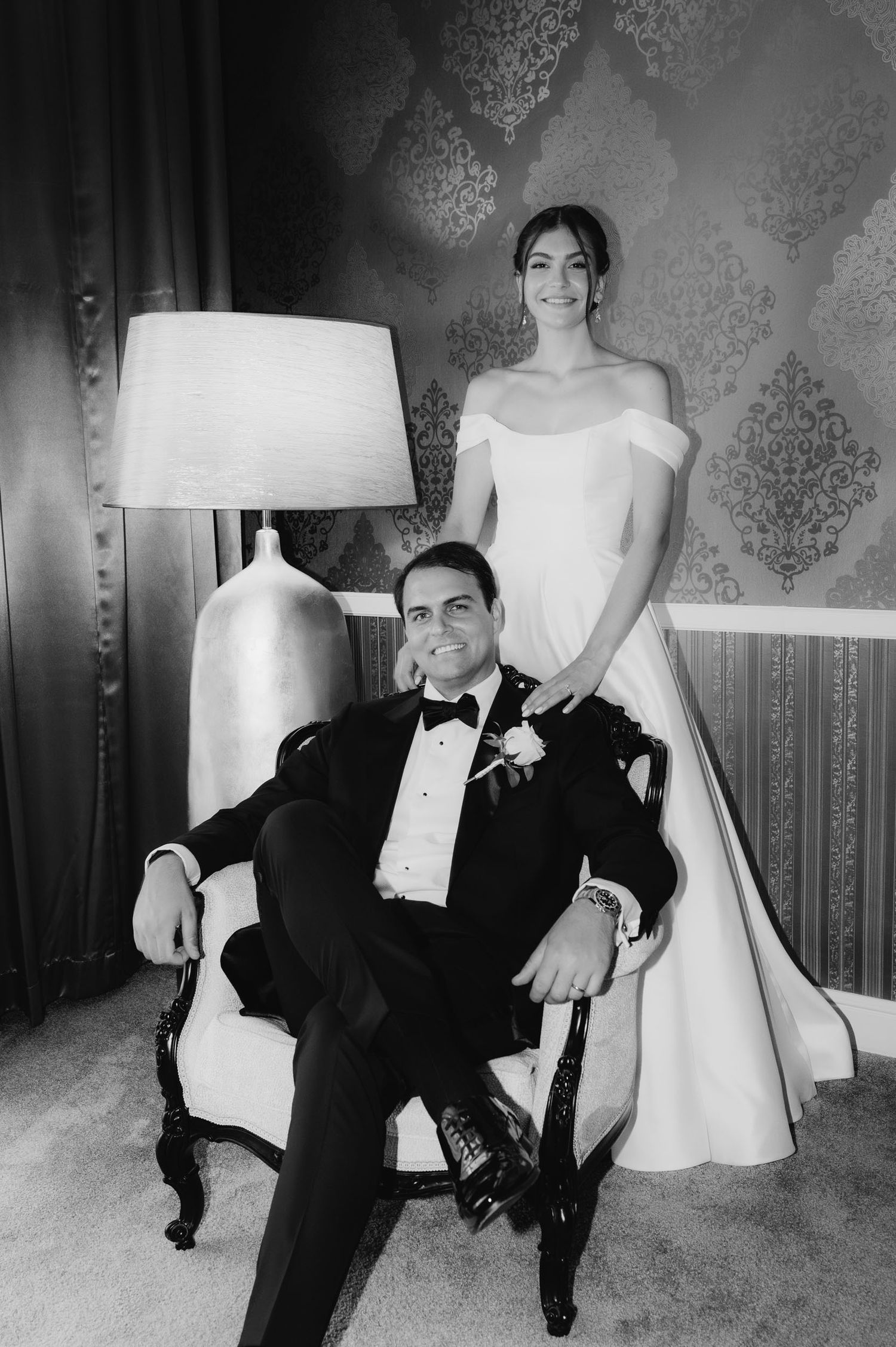 Black and white wedding portrait of bride standing behind groom seated in ornate chair, elegant ballroom setting.