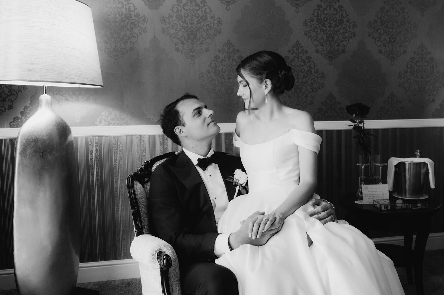 Black and white wedding photo of bride and groom sitting together, smiling at each other in an elegant room.