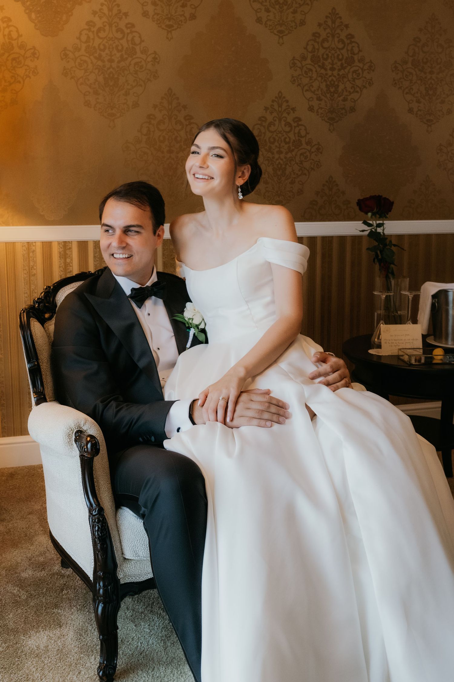Bride in off-shoulder white gown sits on groom's lap in tuxedo, both smiling in elegant gold wallpapered room.